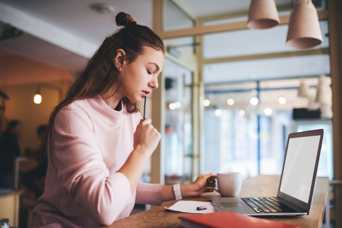 The image is about a laptop and a girl thinking while looking at the paper wile holding a cup of coffee which means checking if you need to file by reviewing income thresholds helps you avoid unnecessary paperwork or missing out on money.
