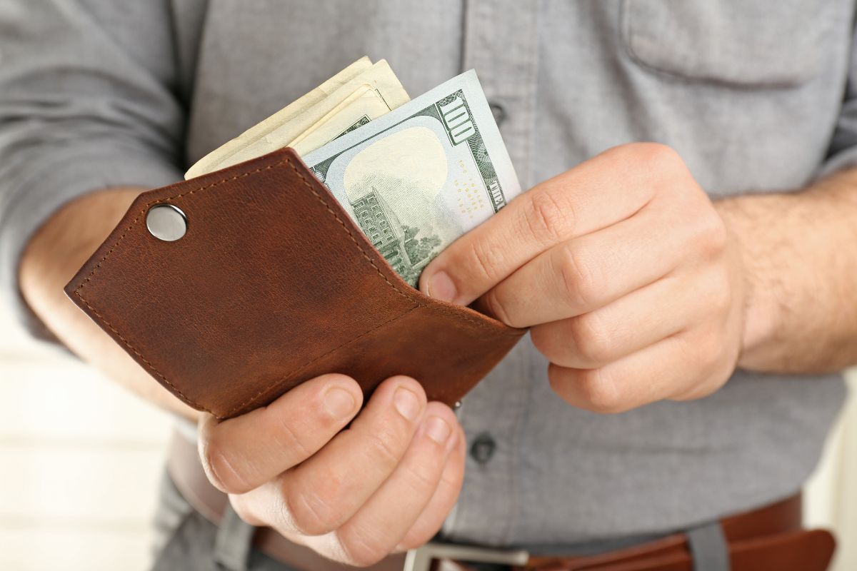 The photo shows a man's hand putting the dollar bills into his wallet referring to setting aside money for self-employment taxes that can prevent a bog tax bill later.