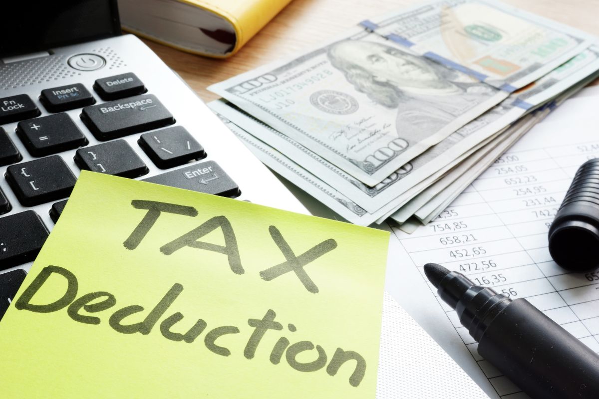 The image shows a laptop, dollar bills, a piece of paper with numbers, a marker, and a piece of paper with notes referring to checking if you qualify for tax deductions that can help reduce your owe or increase your refund.