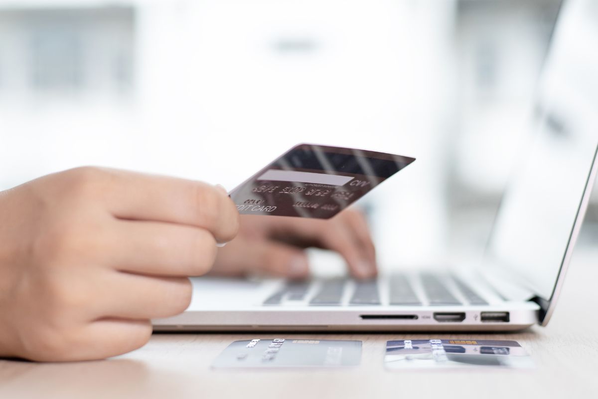 The photo shows a man's hand using a laptop while holding a credit card, indicating that using a secured credit card creatively can help you qualify for regular credit cards.