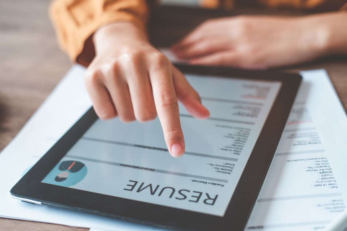 The image shows papers on the table and a girl's hand using a tablet device with a screen display referring to the use of AI resume builders to help fix grammar, make a resume look more professional, and get interviews faster.