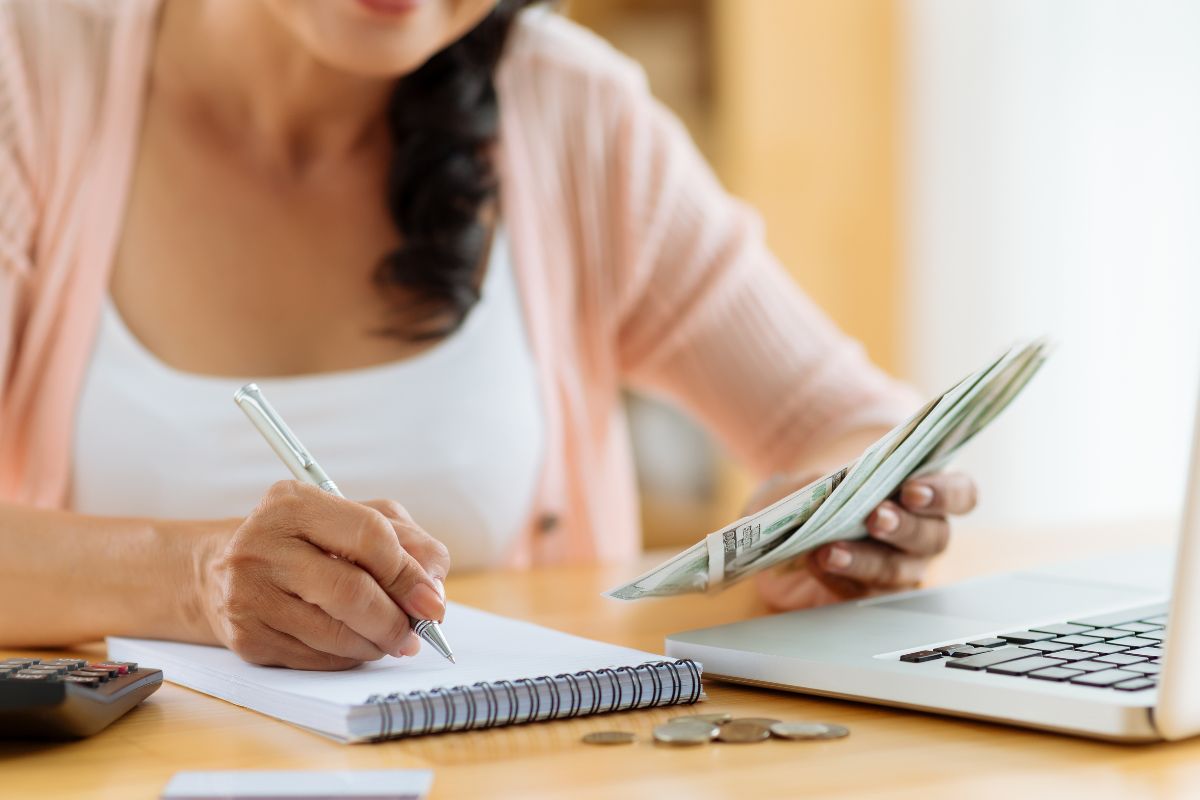 The picture shows a laptop, coins, a calculator, and a girl's hand holding money while writing in the notebook which means to use a reverse budget by covering essentials and savings first to allocate money for future needs and spend what's left.