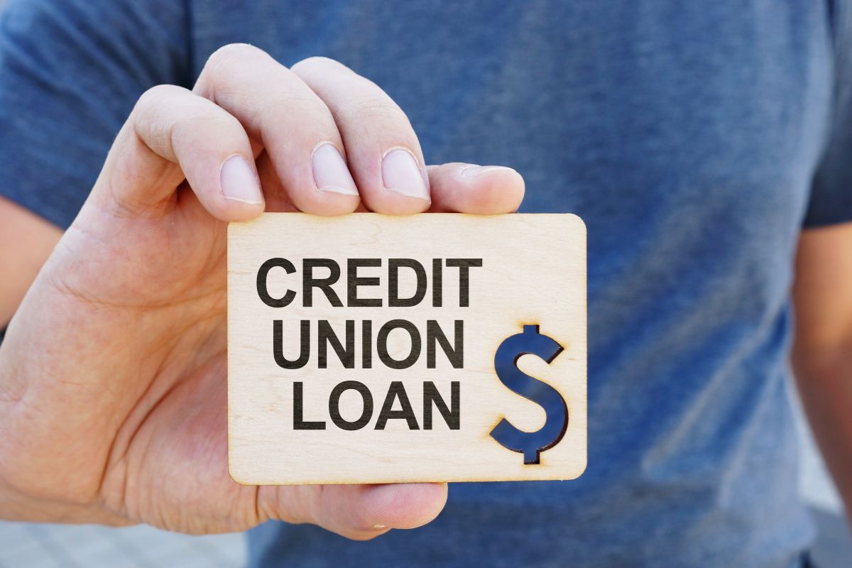 The image shows a man's hand holding a wooden with notes and a dollar sign engraved, which means using a credit-builder loan to help people with low or no credit.
