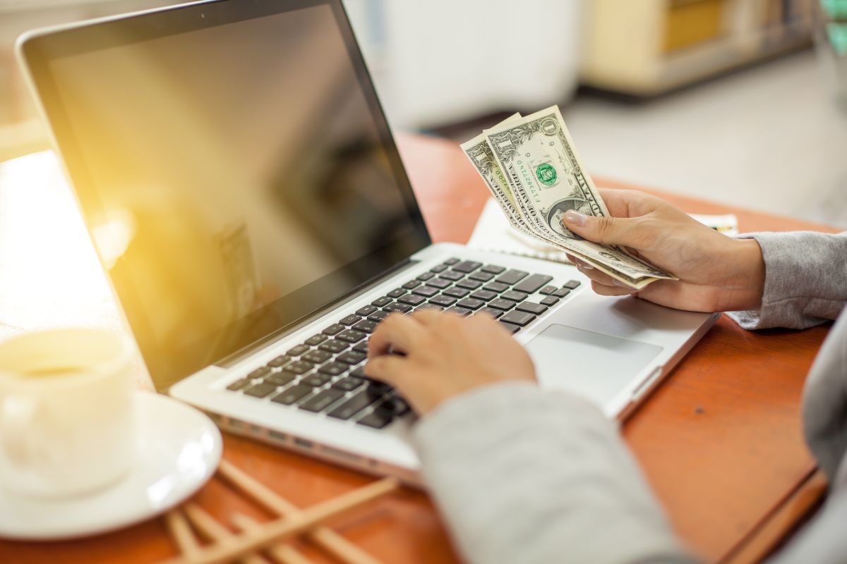 The photo shows a coffee and a girl's hand typing on the keyboard while holding dollar bills, which means setting up autopay with extra payments can cut down interest costs and help your credit score over time.