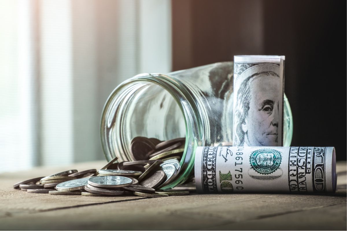 The photo shows rolled dollar bills and a jar with coins which means creating a spending jar and seeing how much small choices add up can make it easier to stick to the challenge.