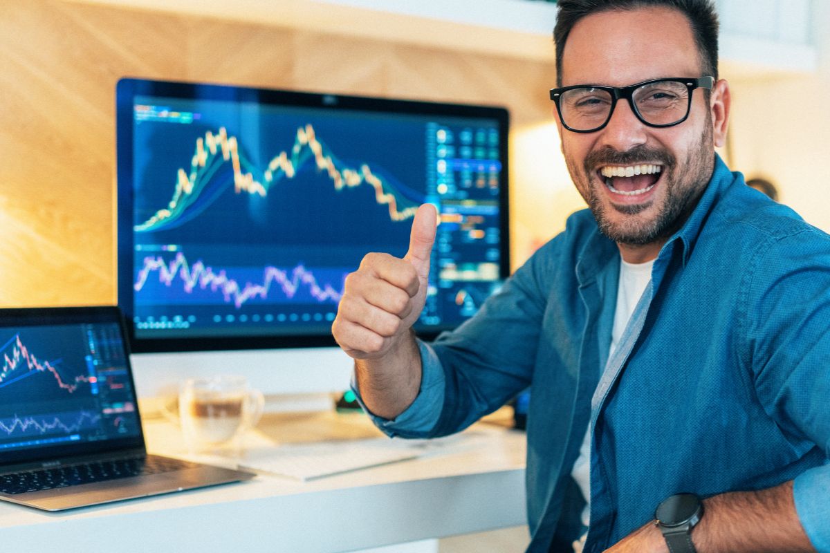 The image tells about a computer and laptop with a screen and a man doing an ok sign display which means a day trader can be a way to make money without needing a traditional job. 