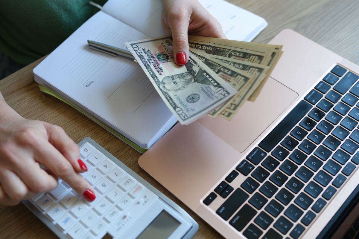 The image shows a laptop, a notebook, a pen, and a girl's hand holding dollar bills while calculating referring to enjoying living below your means that can be put toward investments, business opportunities, and long-term financial freedom.