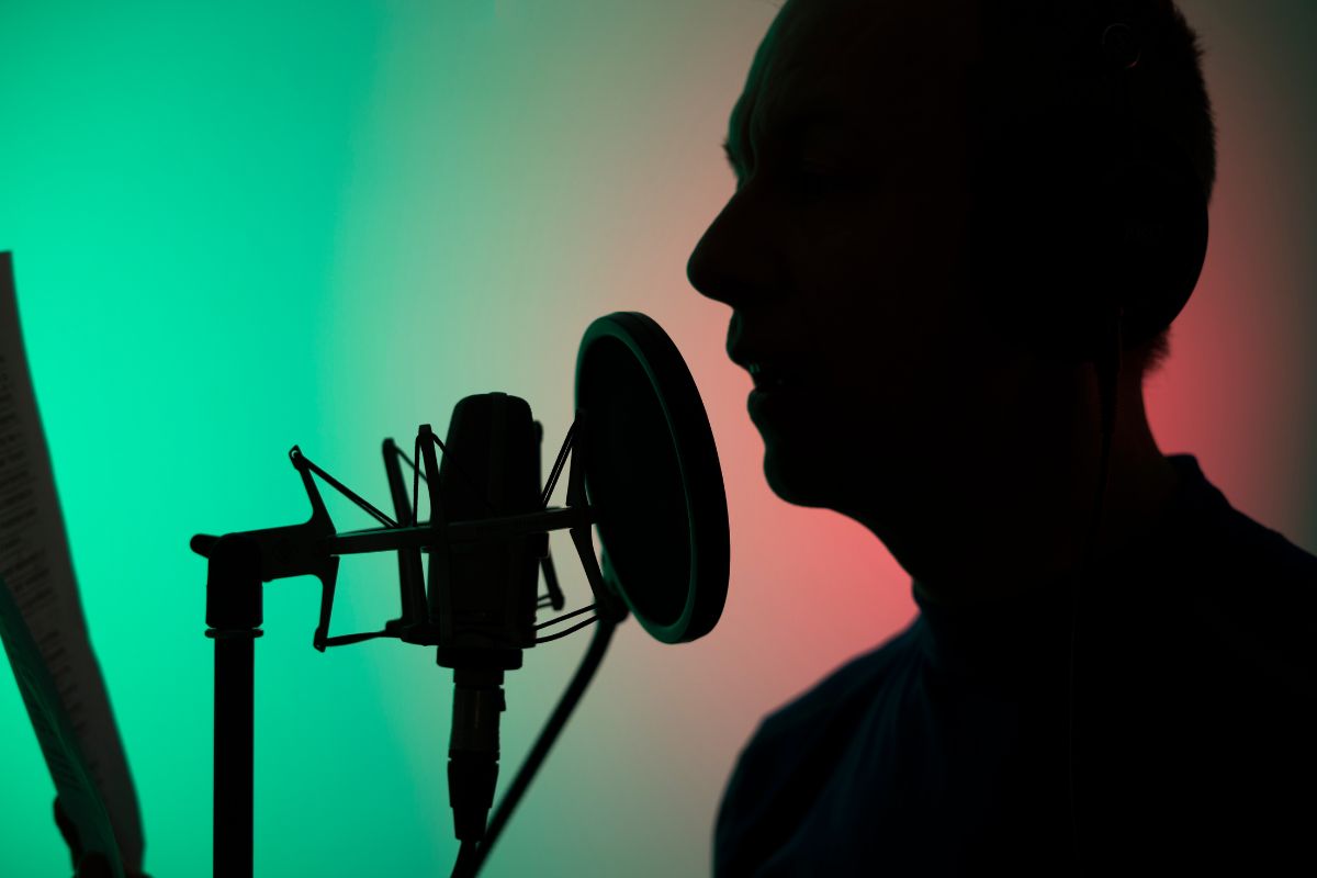 The image shows a man recording his voice using a microphone which means a freelance voiceover artist can make money recording audio for ads, audiobooks, or cartoons and lets you work from home that fit your schedule.