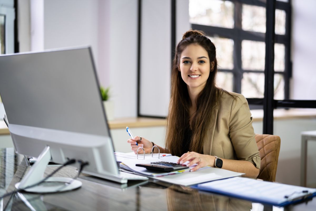 The photo talks about a computer, a clipboard, and papers, and a girl holding a pen while using a calculator indicating that a bookkeeper is a solid part-time job you can do from home and make a steady income.