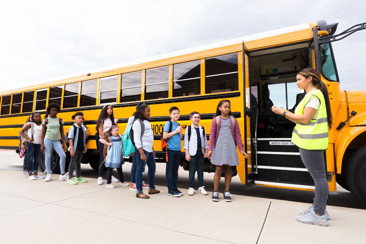 The image shows a girl and students lining beside the bus which means becoming a school bus monitor helps make sure kids are safe while traveling to and from school while still earning steady pay.