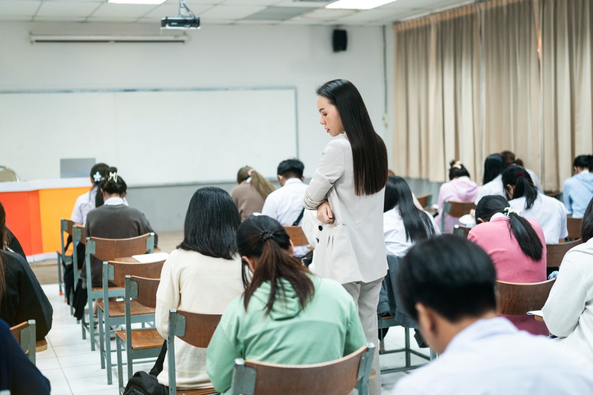 The photo shows students on their exam period and a girl checking on them which means becoming a test proctor is a great way to make extra money without working every day.