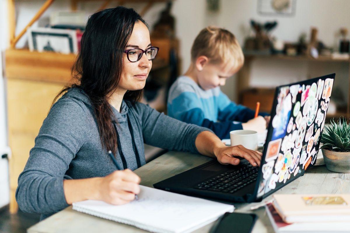 The image tells about a child writing on paper and a girl researching using a laptop while taking down notes indicating to check out the best jobs for moms that let you earn money without taking too much time away from what matters most.