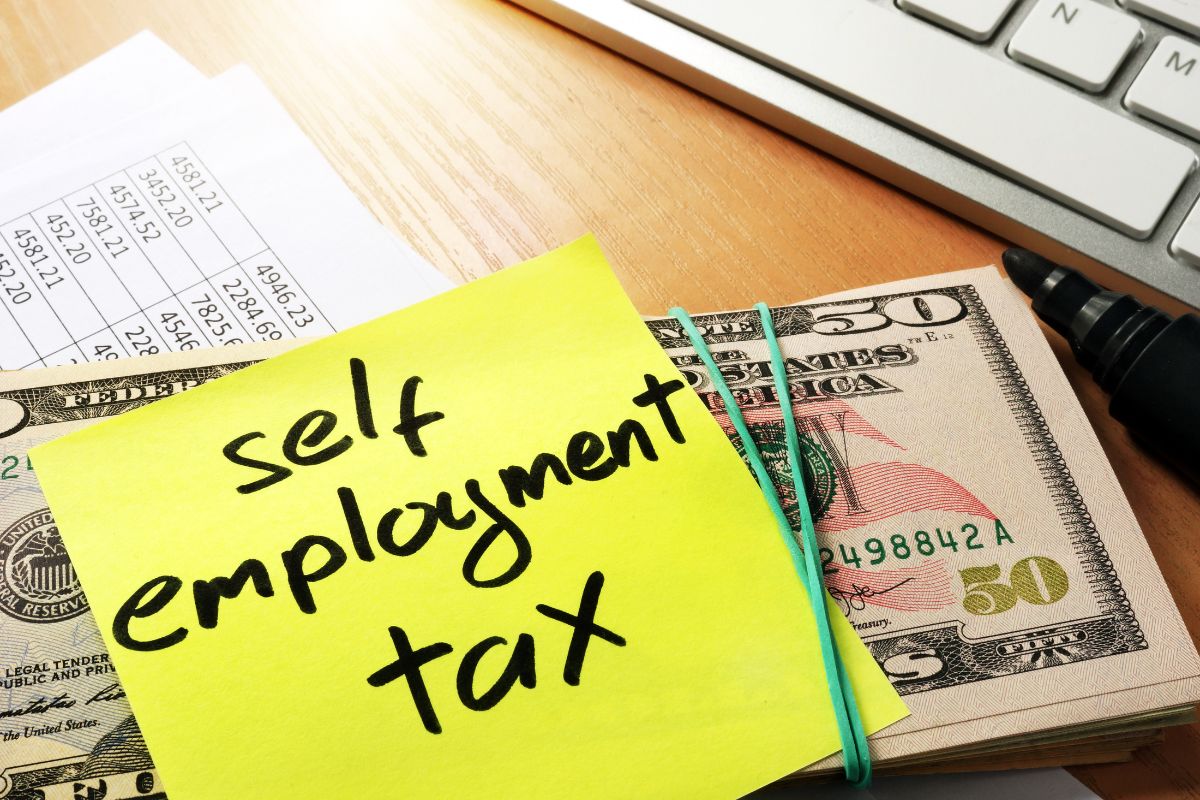 The image shows a keyboard, dollar bills, a marker, papers on the table, and a colored paper with notes indicating that overpaying self-employment taxes means giving away money, right tax breaks can lower the amount owed.