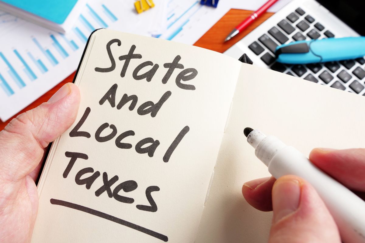 The photo shows a laptop, a pen, a notebook, papers, and a girl's hand and writing on the notebook which means forgetting state and local tax deductions could mean paying more in federal taxes than necessary, checking it can lead to savings.