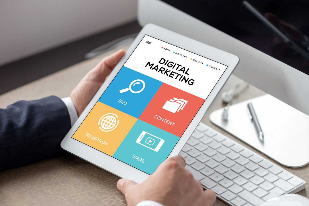 The photo shows a computer set, a pen, and a man's hand holding a tablet device with a screen display which means digital marketing helps growing businesses online and is one of the in-demand skills to learn and earn money while working with passion.