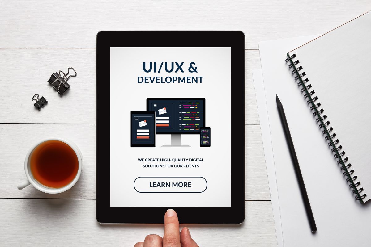 The picture tells about a notebook, a pen, clips, a tea, and a man's hand pointing the tablet device with a screen display indicating that HUX/UI Design is an in-demand skill for designing a user-friendly website and digital experiences.