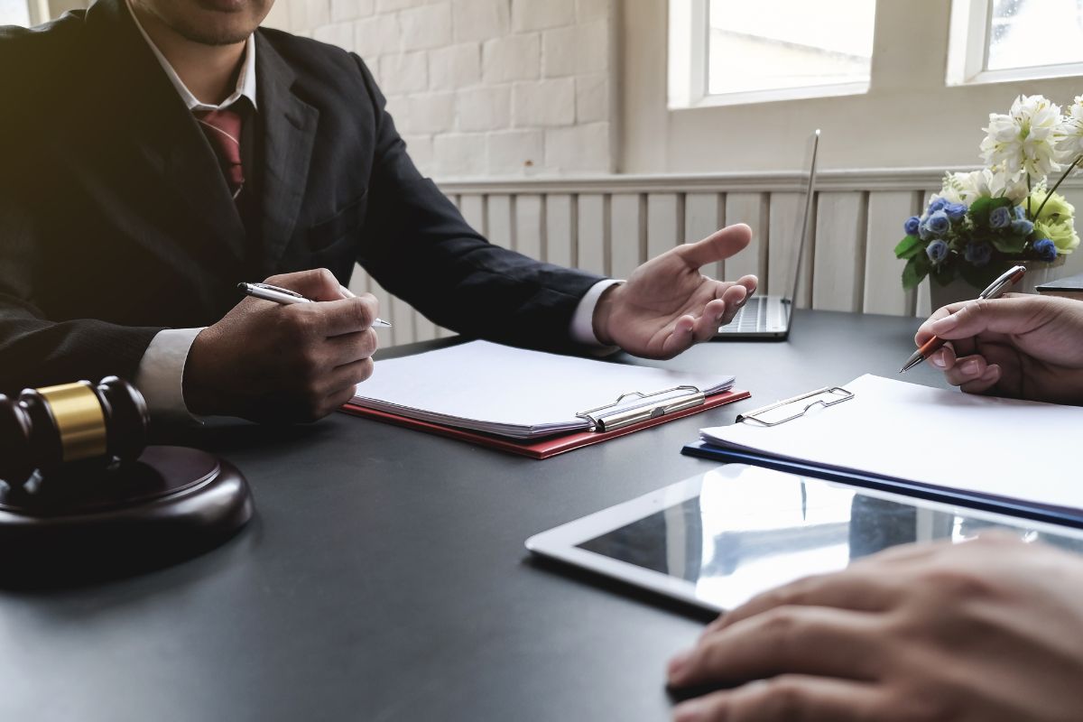 The picture shows a gavel, a laptop, and a man with a clipboard explaining to the man with a tablet and taking down notes indicating that legal and compliance consulting helps clients guide them with business regulations and with risk management.