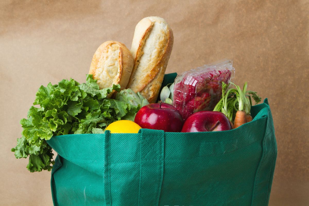 The picture shows a reusable bag with fruits, vegetables, and breads inside which means bringing your bags for discounts to keep more money in your pocket while reducing waste.
