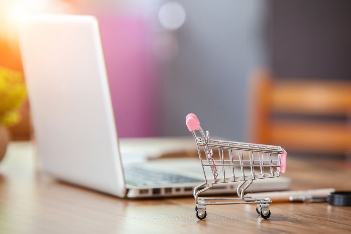 The picture shows a laptop and a mini cart on the table which means learning more tricks to save money on groceries can keep more money in your pocket and find the best deals.