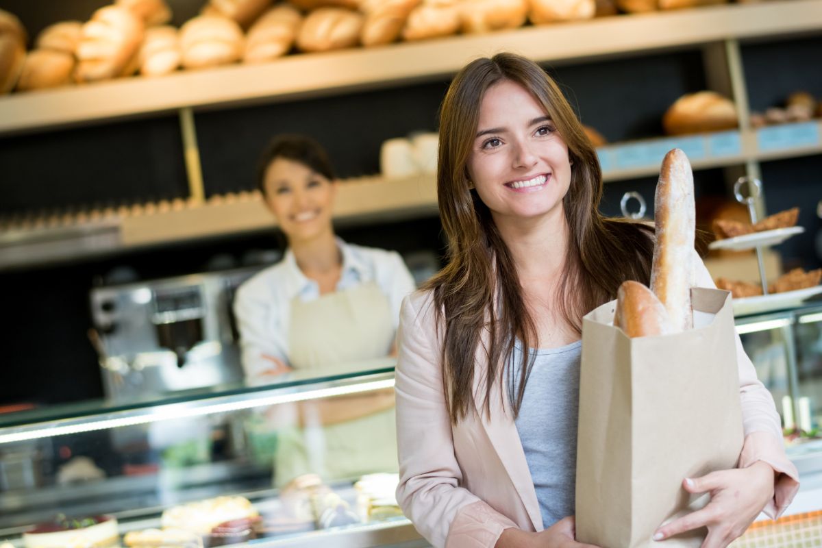 The photo tells about breads on display, a cashier, and a girl carrying a paper bag with bread inside which means to shop at closing time for discounts is an easy way to save money.