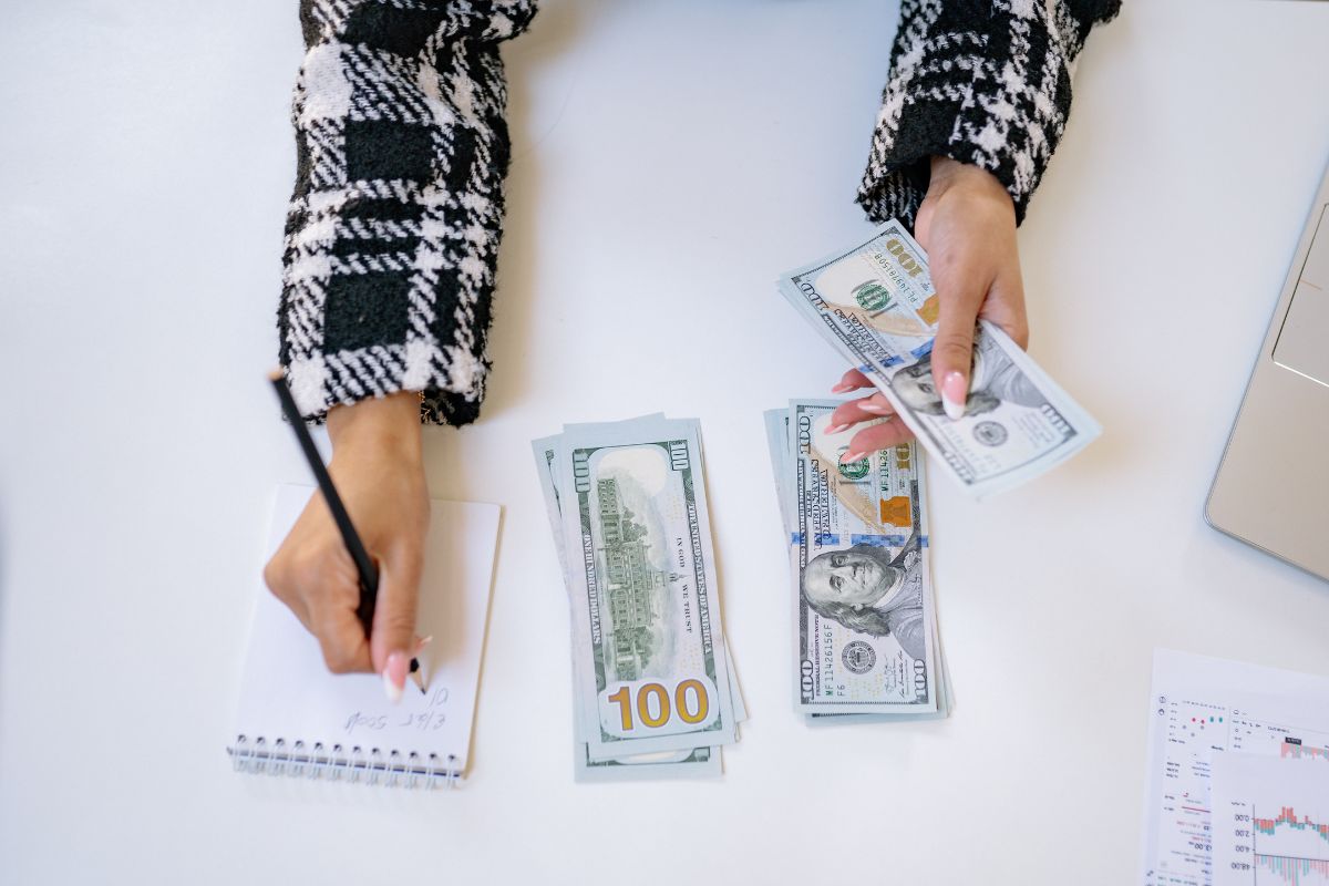 The picture shows dollar bills on the table and a girl's hand writing on the notebook while holding dollar bills which means tracking evert dollar keeps from wasting money and helps make smart financial choices.