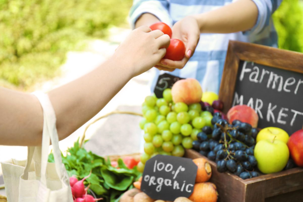 The photo shows a woman buying vegetables at a farmer's market.