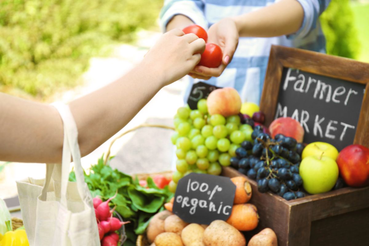 The photo shows fruits with notes on boards, and a customer's hand handing the tomatoes to the vendor's hand referring to living below their means which gives extra money to invest and save.