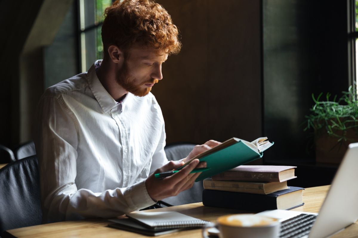 The picture of books, a laptop, a coffee, notebooks, and a man reading them indicating to read financial news or books helps make smarter choices and get at growing your net worth.