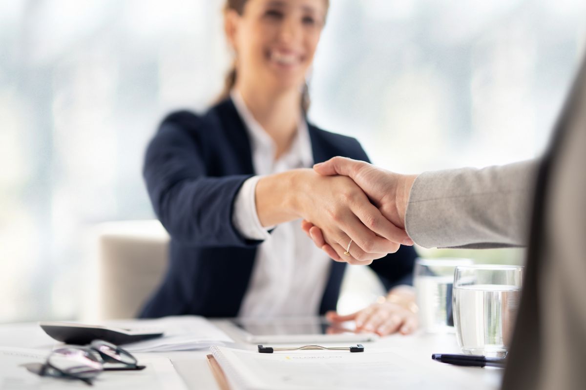 The picture shows papers on the table, water, a calculator, an eyeglass, and two people shaking hands together which means negotiating everything that adds up over time and saves money.
