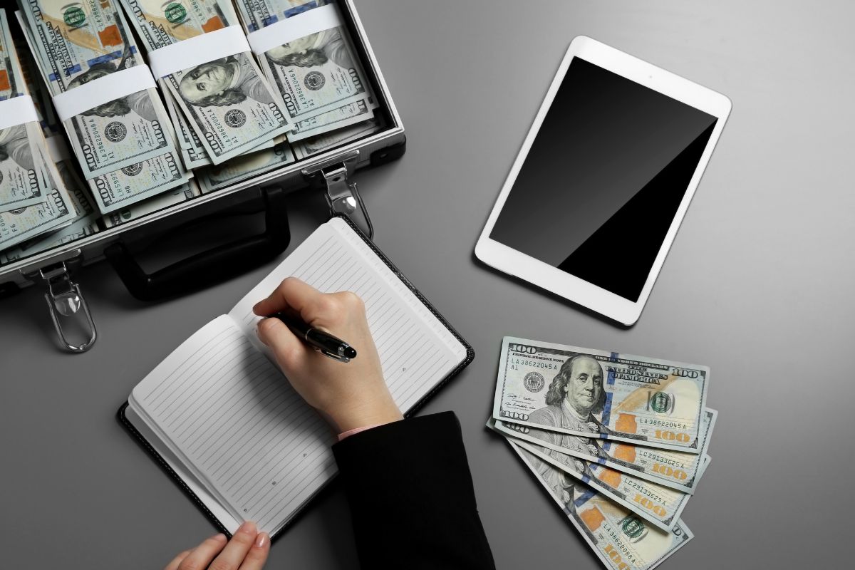 The picture shows a case with dollar bills, a tablet device, dollar bills, on the table, and a girl's hand and writing on the notebook which means to become independently wealthy with these guides helps you reach goals and save more money.