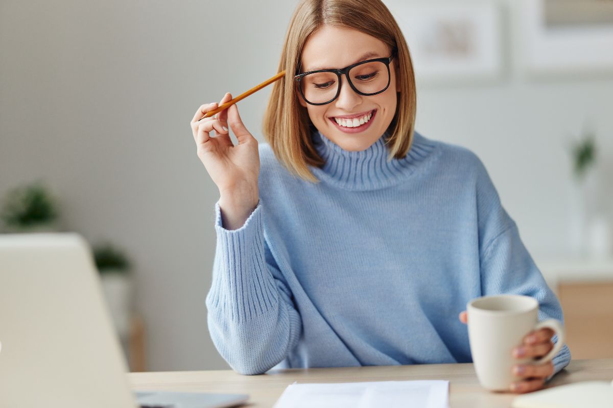 The picture shows a laptop and a girl reading a paper with a coffee and pen on her head referring to creating a money mantra and repeating it before spending can stop you from buying things you don't need and help to make smart choices.