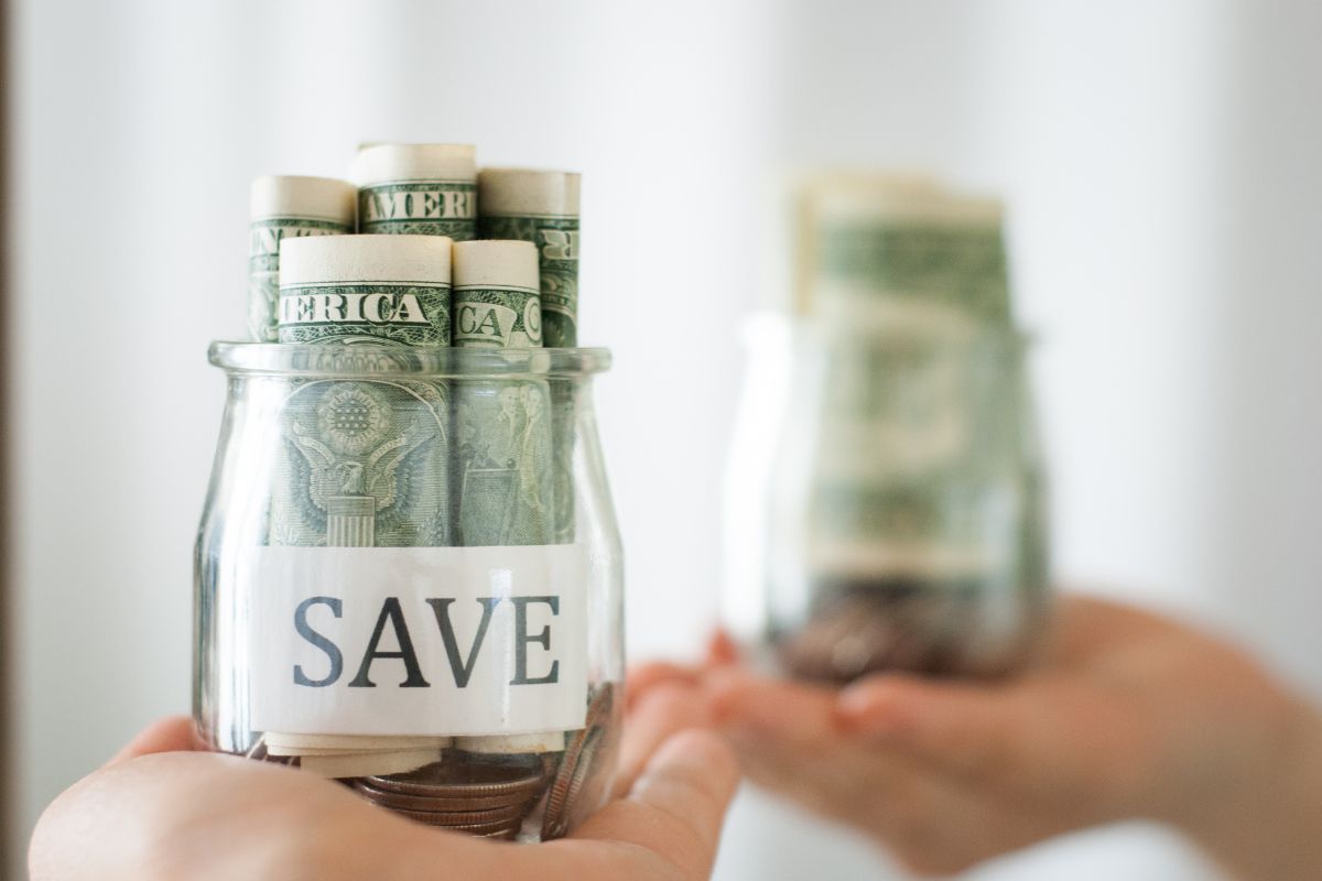 The picture shows a girl's hand holding a jar with coins and dollar bills facing the mirror referring to automate savings in unpredictable accounts is an easy way to build a strong habit without feeling the pinch.