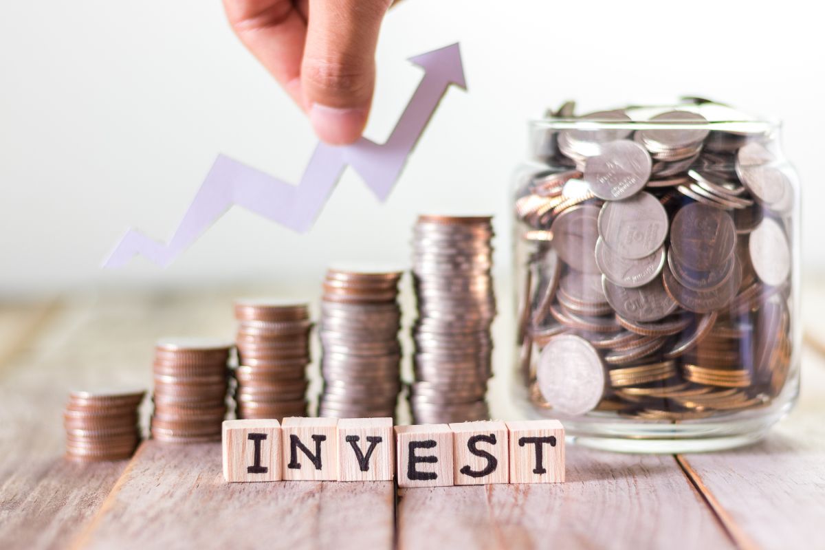 The image shows a jar filled with coins, coin stocks, wooden cubes with letters, and a man's hand holding an arrow referring to the focus on daily wealth-building actions to help build wealth in the long run.