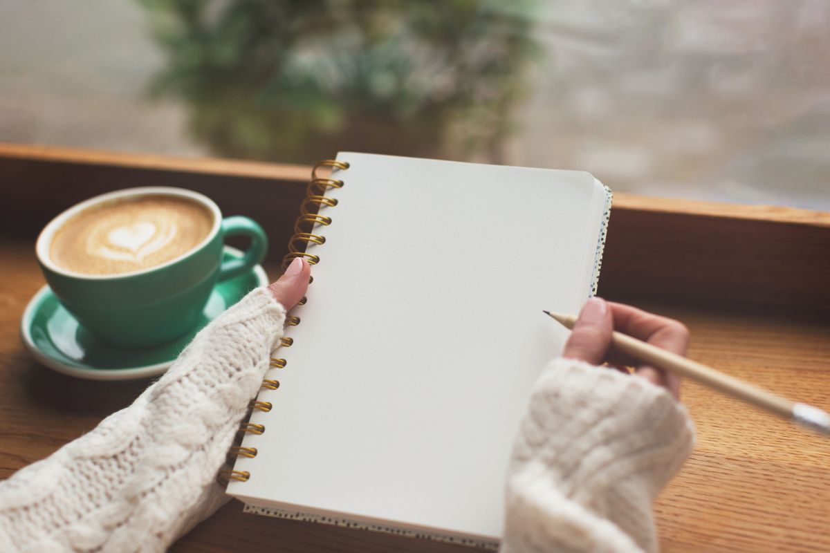 The image shows a coffee and a girl's hand holding a notebook and a pen indicating that writing your financial goals helps you act in ways that support your goals and make smart choices with money.