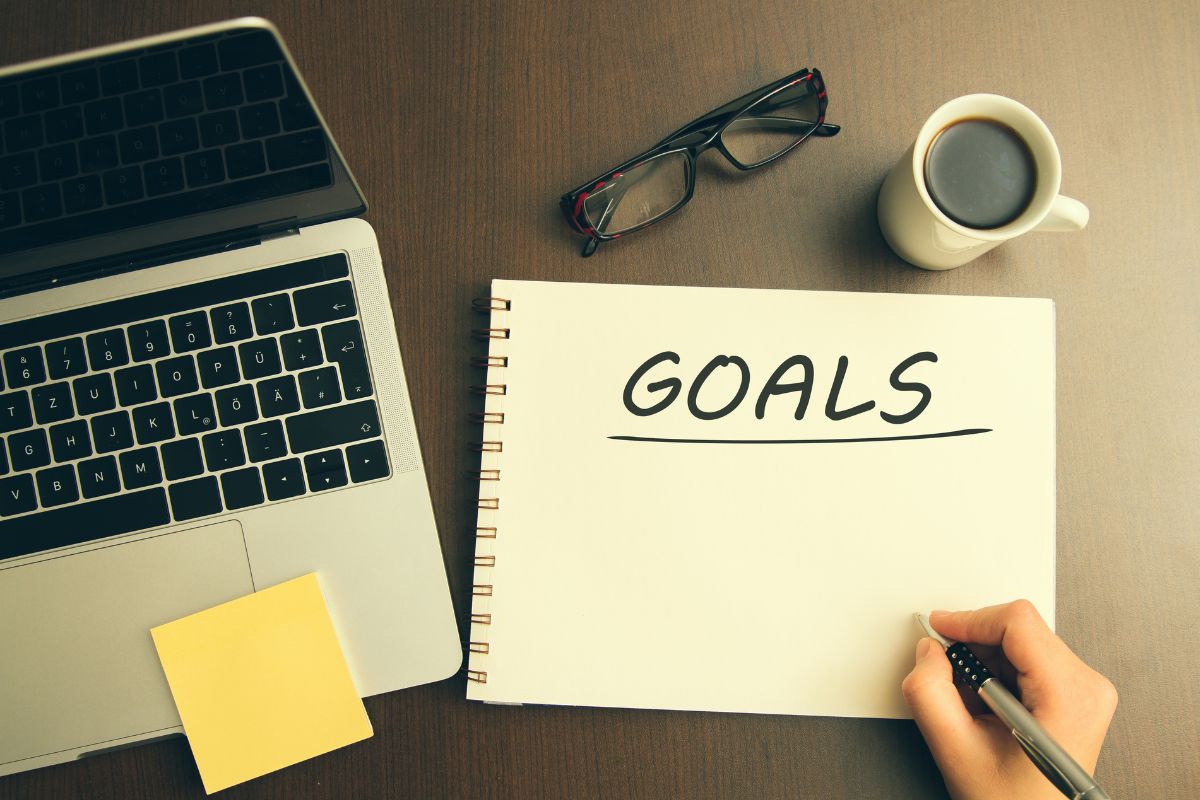 The picture shows a laptop, an eyeglass, coffee, and notebook with a note, and a girl's hand holding a pen referring to the use of the domino effect strategy builds confidence and motivation to tackle bigger steps.