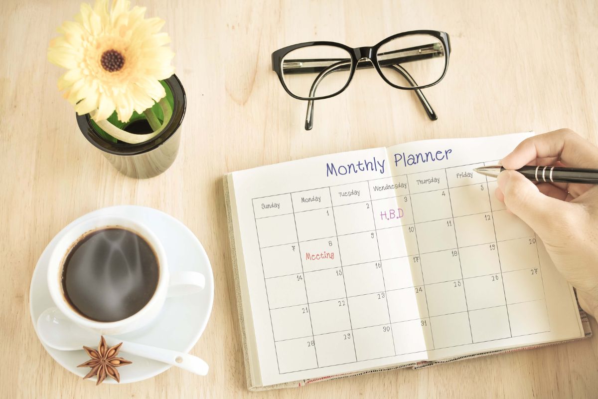The photo shows an eyeglass, a coffee, a flower, and a girl's hand and writing on the notebook with notes which means assigning a theme to each month keeps financial goals fresh and prevents burnout.