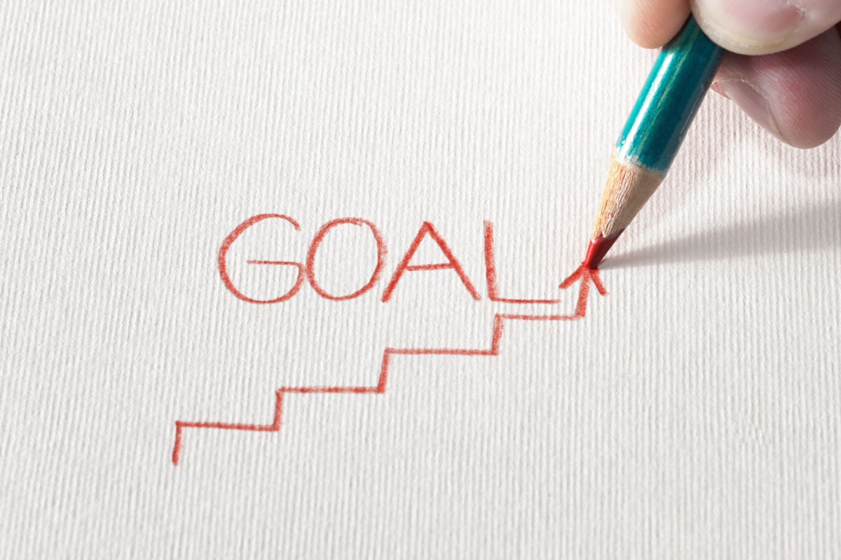 The photo shows a man's hand and writing on the canvas referring to setting stretch goals beyond your main goal puts you in a stronger financial position later.
