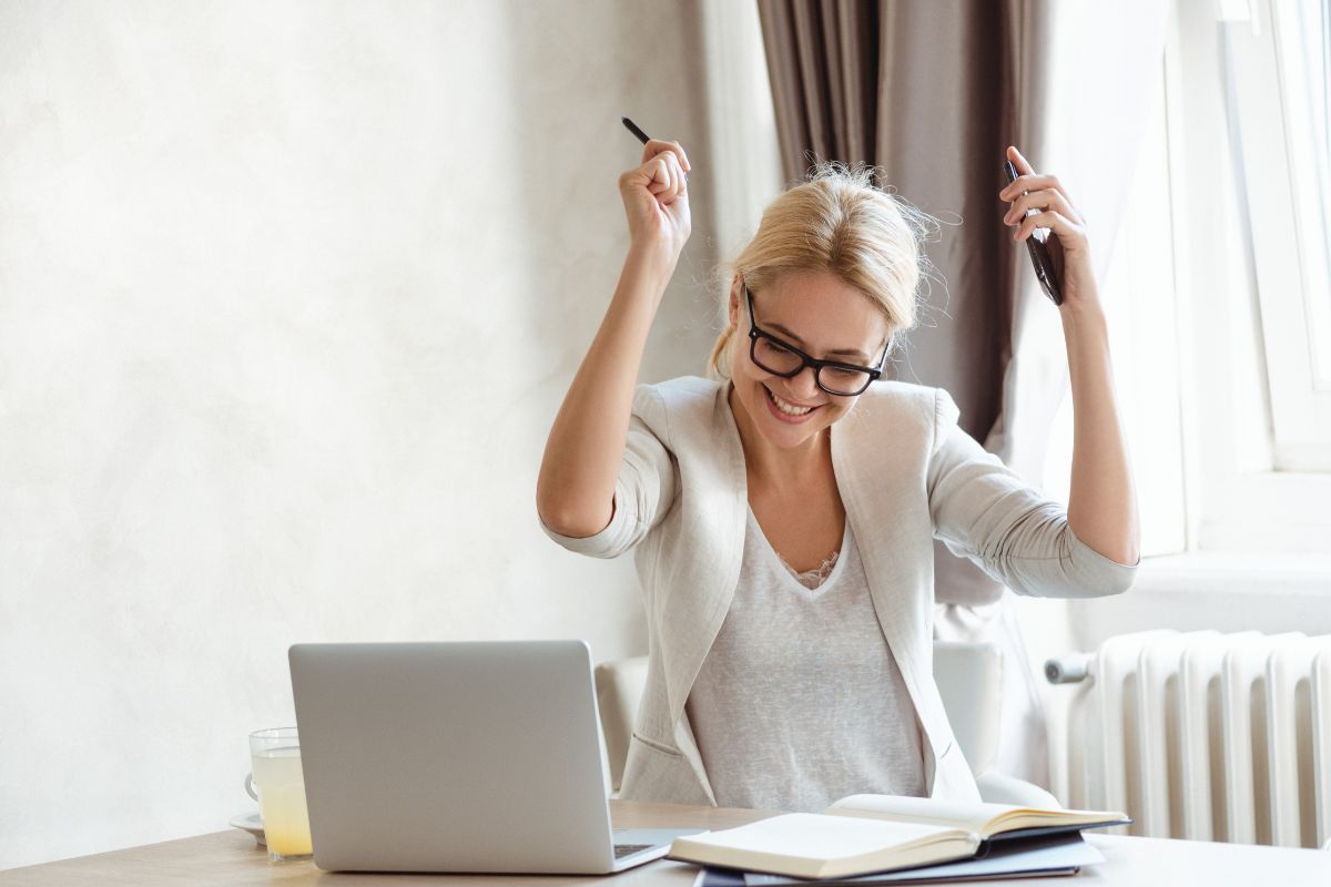 The image shows a notebook, a laptop, and a girl cheering which means tracking every win no matter how small keeps you motivated and keeps going.
