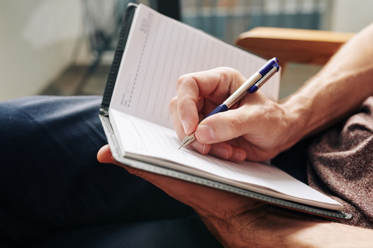 The image tells about a man's hand and writing on the notebook which means to use of pain and pleasure makes financial goals easier to stick with.