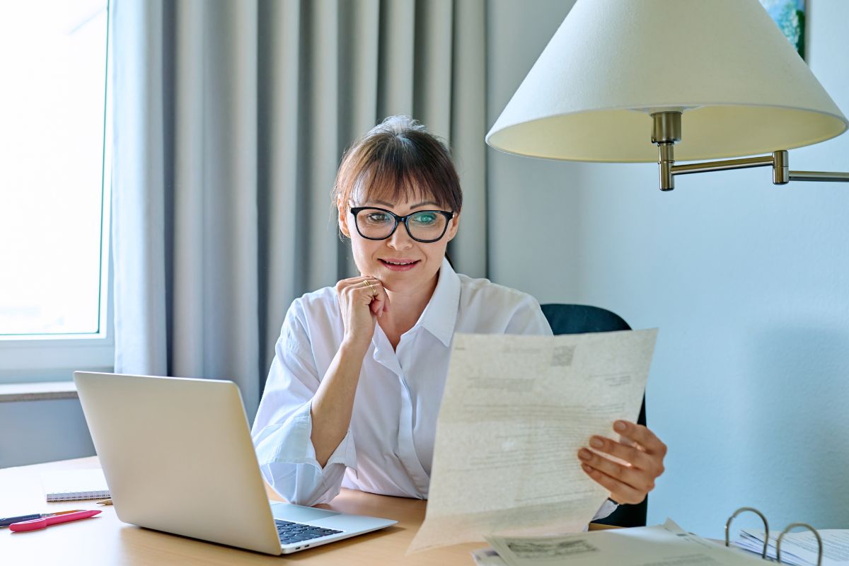 The photo tells about a laptop, papers on the table, and a girl holding a paper and reading it which means keep iterating keeps you moving forward without feeling stuck.