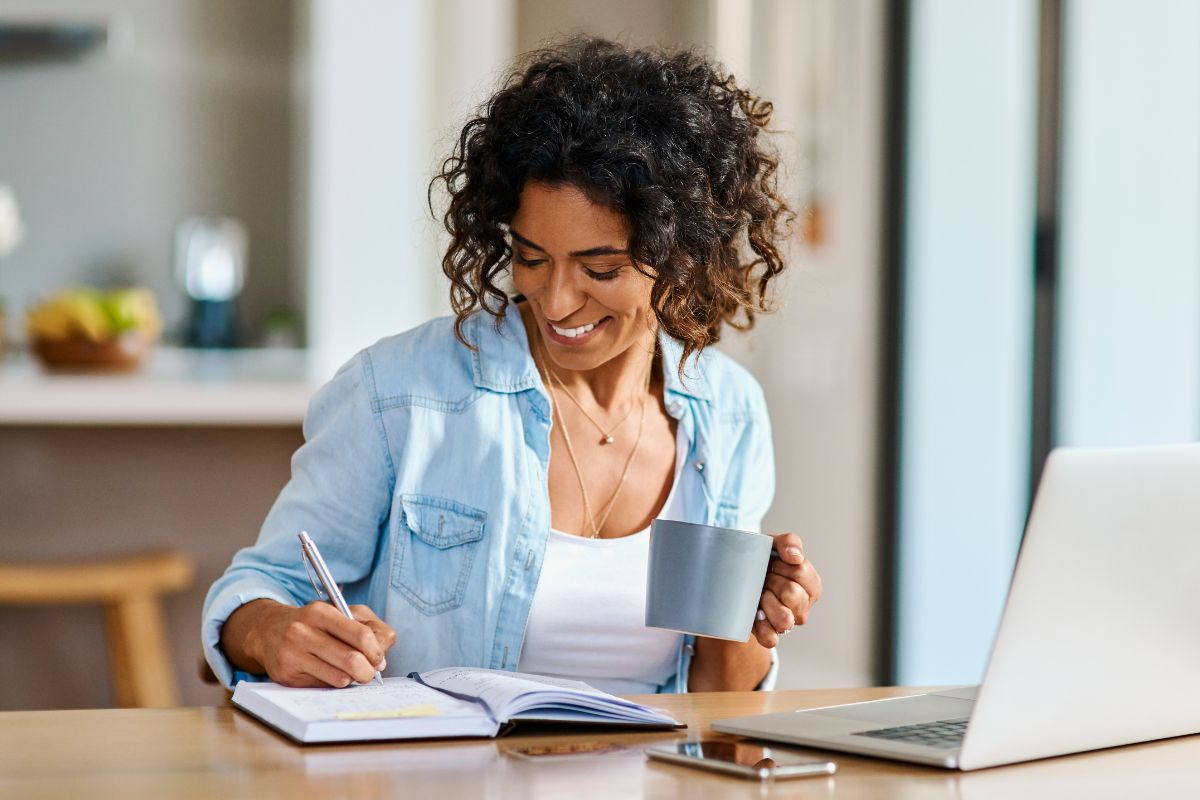 The photo of a laptop, a phone, and a girl writing in the notebook while having a coffee which means writing down career wins every day keeps you motivated and reminds you how far you've come.