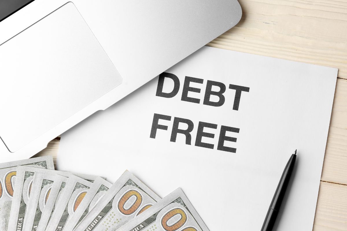 The photo shows a laptop, a pen, dollar bills, and a piece of paper with notes indicating that setting a bold and specific debt-free goal keeps you focused and motivated and easier to stay on track.