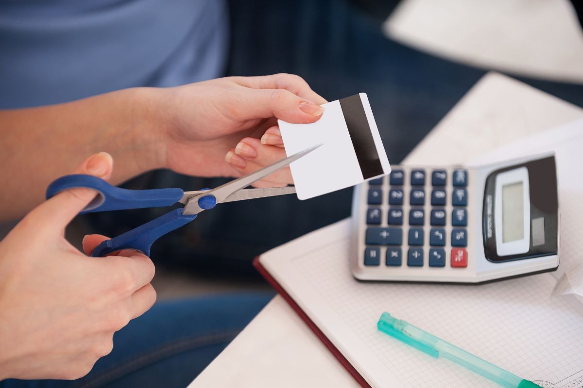 The image shows a calculator, a pen, a notebook, and a girl's hand cutting the card indicating to cut one major expense for a month to lessen the money spent and add up to savings.