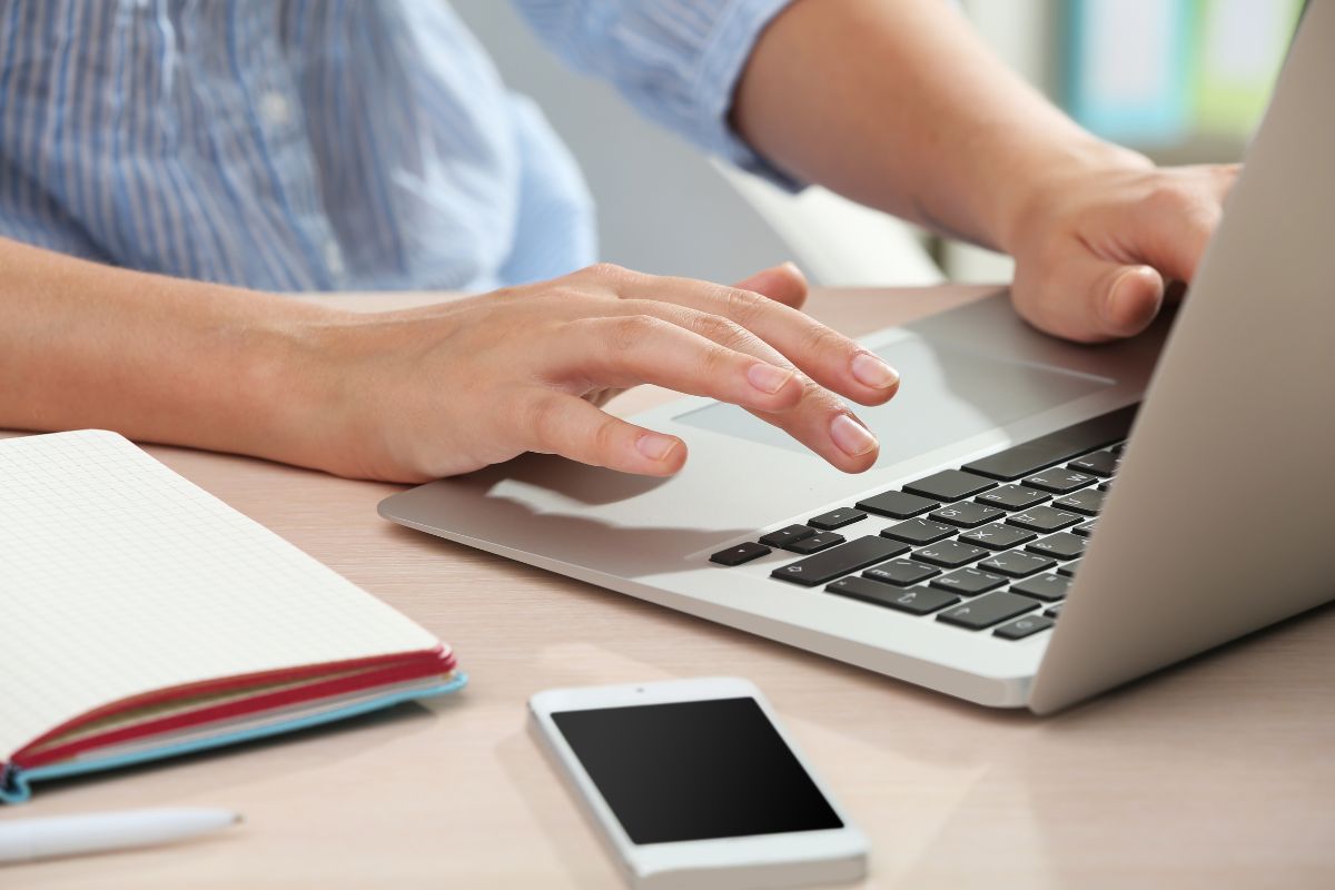 The image shows a phone, a notebook, a pen, and a man's hand using a laptop to find ways to make $500 fast with these guides to reach financial goals.