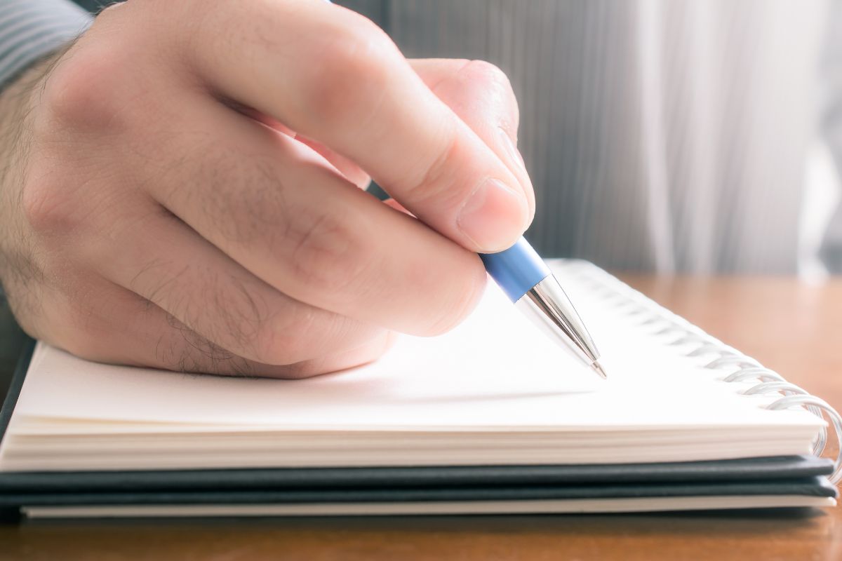 The image shows a man's hand and writing on the notebook which means writing off business expenses if you're self-employed making filing easier and ensuring all possible deductions are claimed.