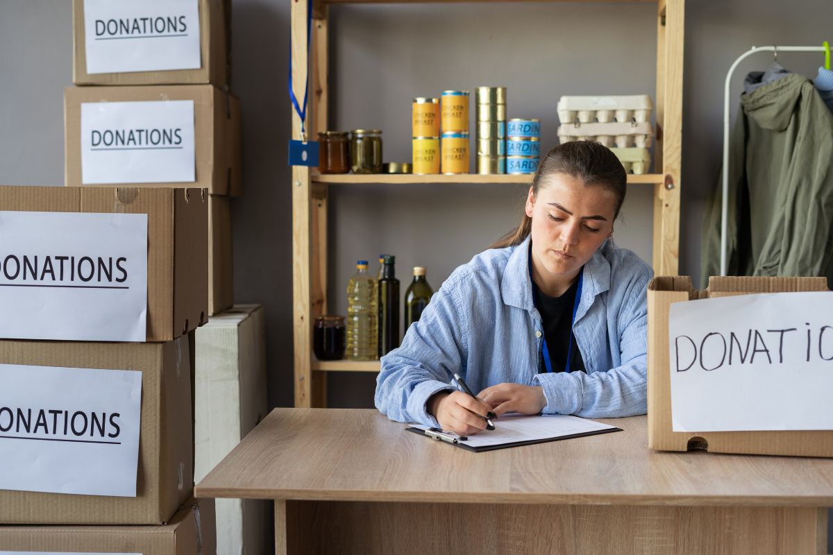 The photo shows boxes with notes, canned foods, and a girl writing on the paper referring to track and deduct eligible charitable donations can lower taxable income and reduce taxes owed.