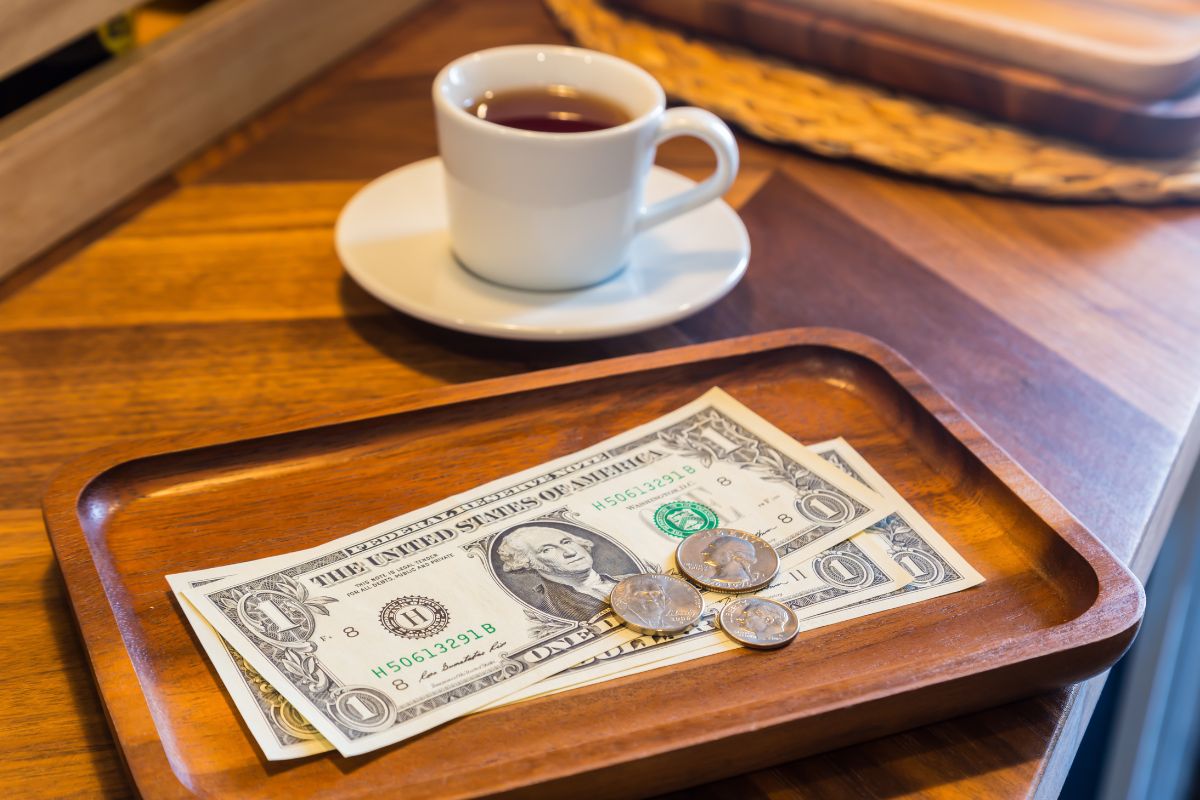 The picture tells about a cup of tea and a wooden plate with coins and dollar bills which means paying tips at restaurants with cash ensures your server gets the full amount.