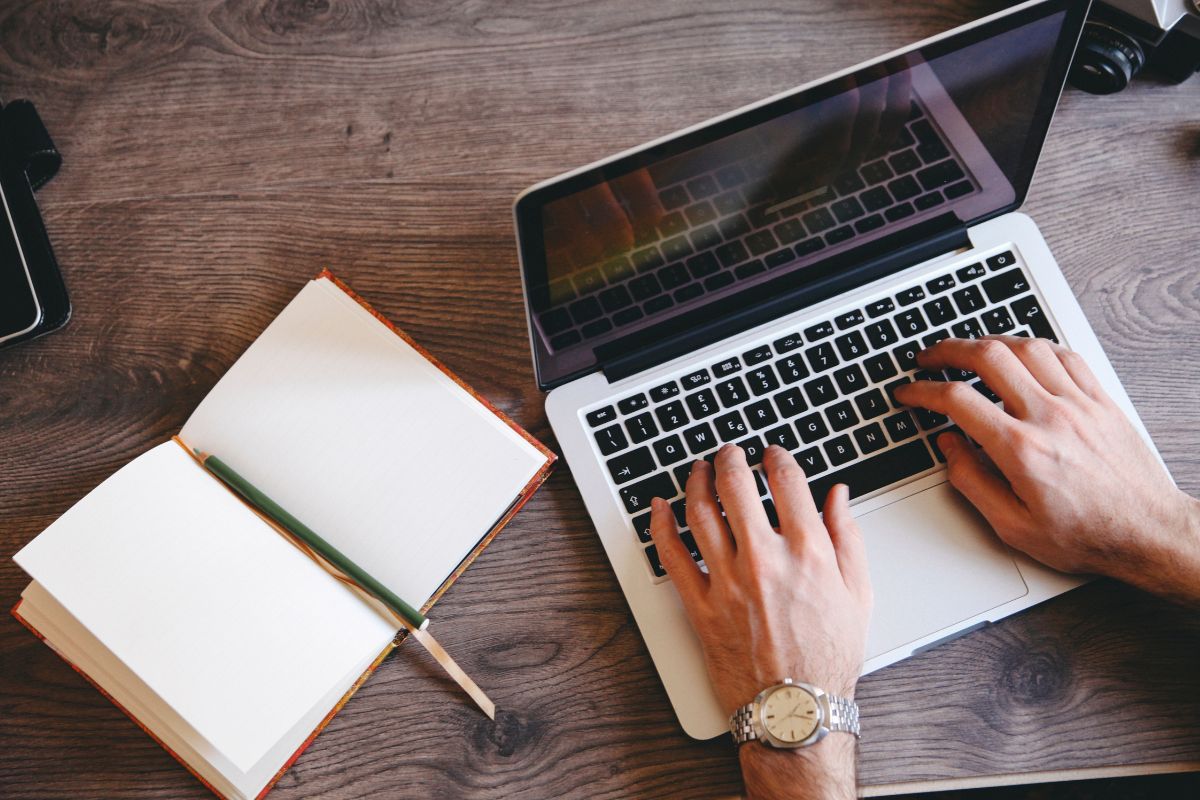 The image shows a notebook with a pen and a man's hand typing on the laptop which means to offer copywriting services that can be a profitable way to make money from home.