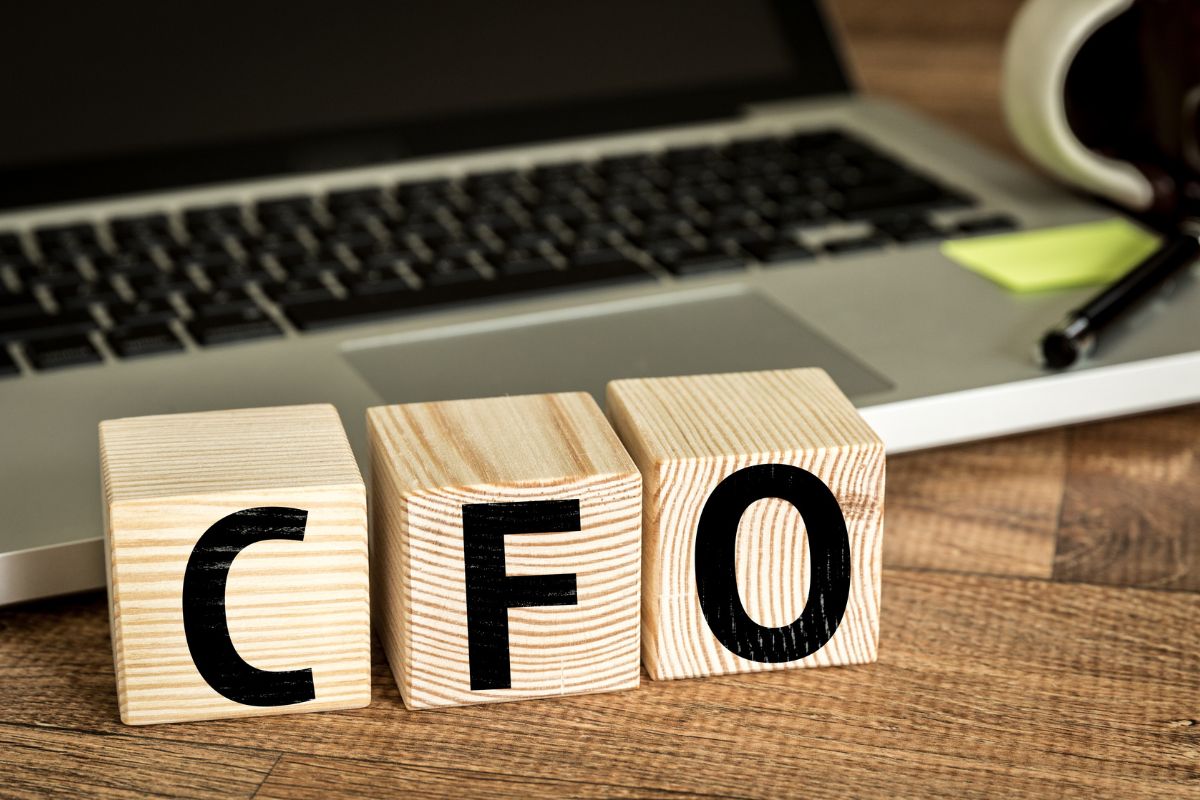 The photo tells about a laptop, a pen, and wooden cubes with letters referring to becoming a virtual CFO for entrepreneurs is a steady way to make money online and help business owners understand their numbers.