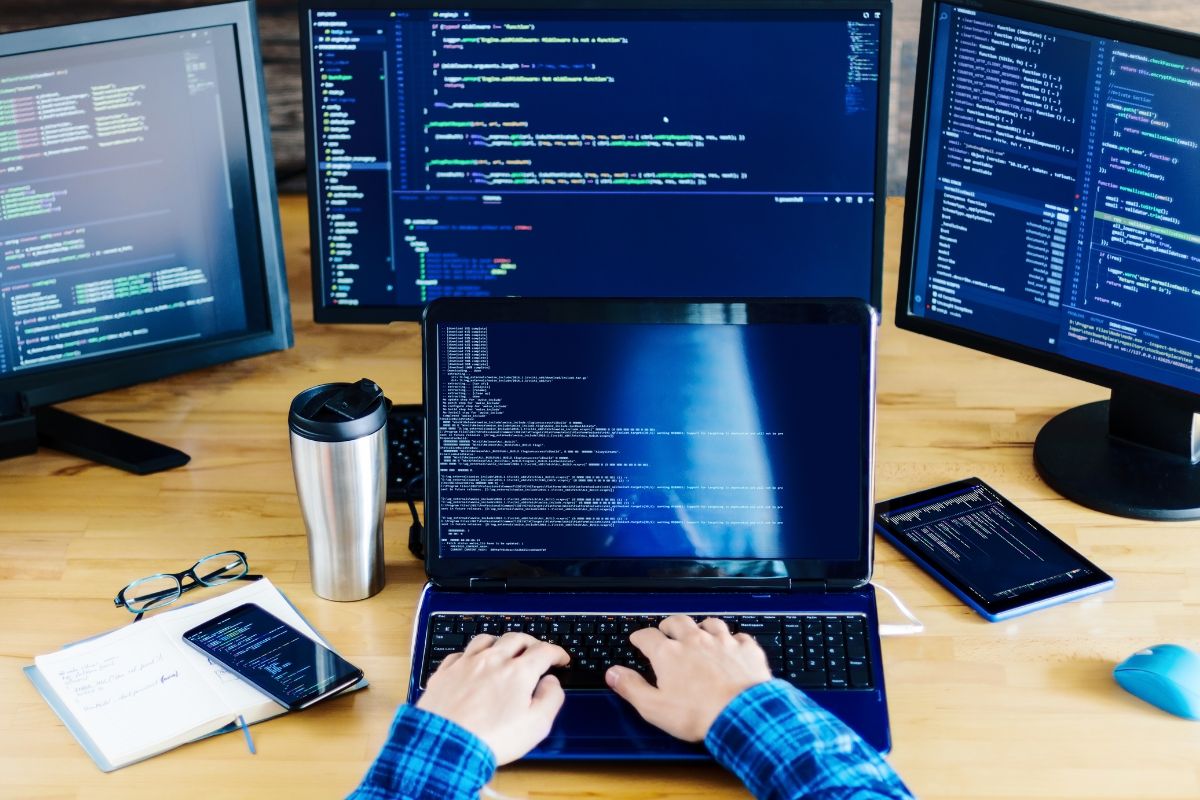 The photo shows monitors, a notebook, an eyeglass, a tumbler, a tablet device, and a man's hand coding on a laptop which means developing and selling niche software can be a great way to make money online. 